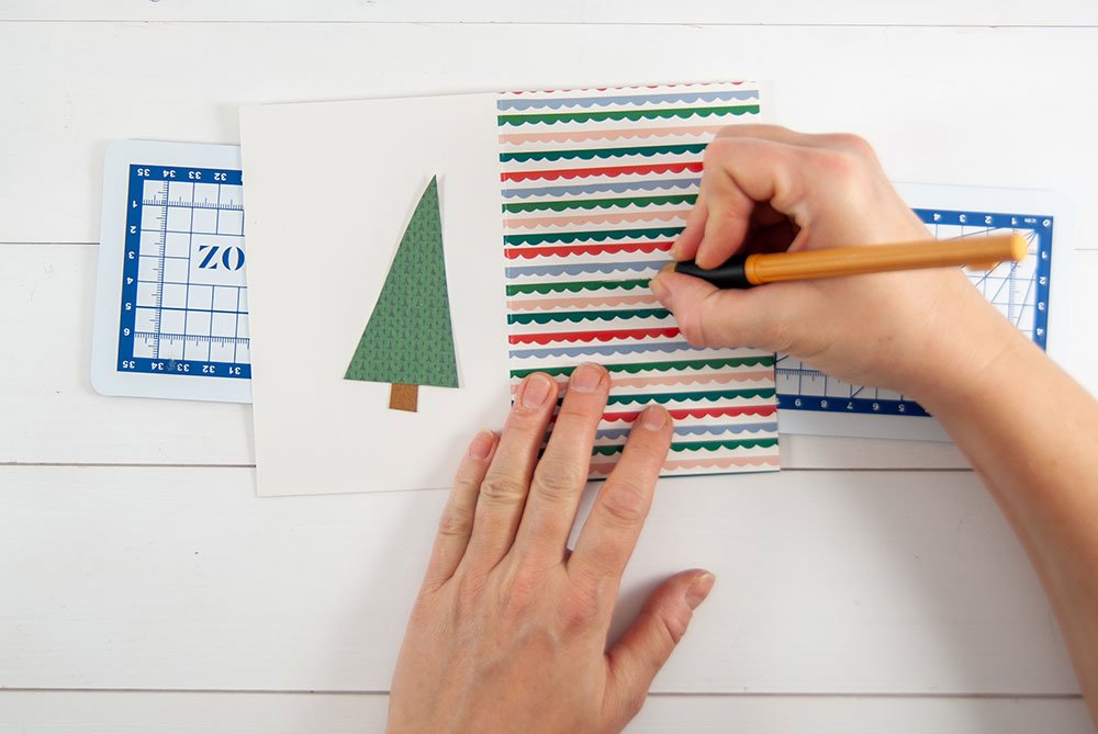 Auf einer blau-weißen Schneidmatte von Artoz wird eine Motivkarte gebastelt.  Eine Hand hält ein Cuttermesser, mit welchem ein Schlitz in eine Karte geschnitten wird. 