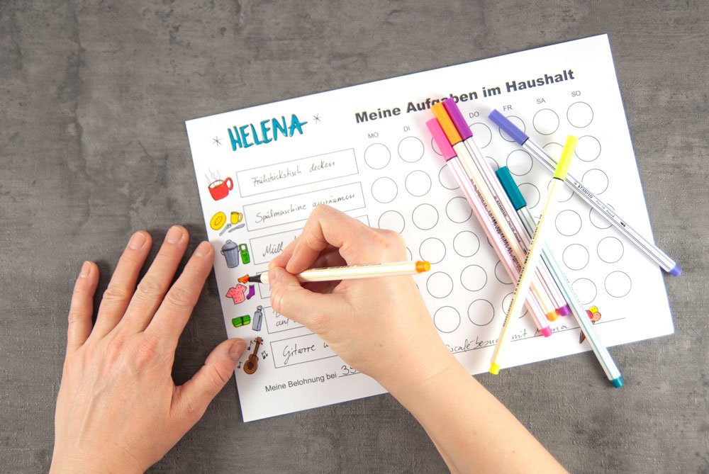 Ein Kind malt die Zeichnungen für jede Aufgabe auf der Aufgabentafel. Viele bunte Stifte liegen auf dem Plan.