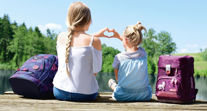 Zwei Mädchen sitzen auf einem Steg vor einem See. Neben ihnen stehen ein Step by Step Schulranezen und ein coocazoo Rucksack. Die beiden Mädchen bilden mit ihren Händen ein Herz.