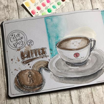 In das Toned Watercolour Book von Hahnemühle wurde mit Aquarellfarben eine Tasse Kaffee und Kekse gemalt. Das Buch liegt auf einem Holzboden. Im oberen Bildrand liegt ein Papier mit verschiedenen Farbproben der Aquarellfarben.