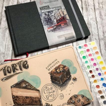 Auf einem Holzboden liegen ein geschlossenes Toned Watercolour Book von Hahnemühle in schwarz. Darunter liegt ein geöffnetes Buch mit cremefarbenen Papier, in das mit Aquarellfarben verschiedene Kuchen gemalt wurden. Rechts davon liegt ein Blatt mit verschiedenen Farbproben von Aquarell-Farben. 