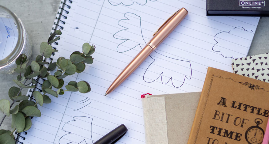 Notizbuch mit Zeichnungen, zwei Stiften und Deko-Elementen auf einem Tisch.