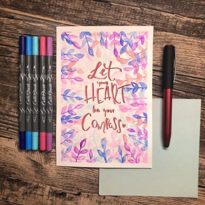 Eine hangeletterte Karte in Lila- und Blautönen liegt zusammen mit Brushpens auf einem Holztisch.