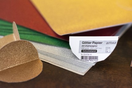 Selbstklebendes Glitzerpapier der Marke Artoz in gelb, grün, rot, gold und silber liegen auf einem Holztisch. 