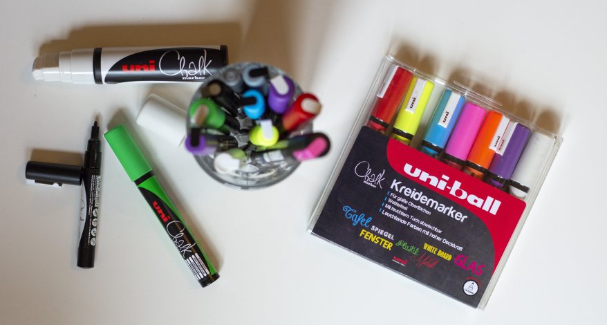 Auf einem Tisch liegen Kreisemarker in unterschiedlichen Farbe der Marke Uniball. Einige Kreisemarker stehen in einem Glas daneben. 