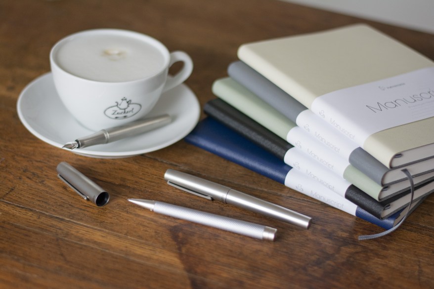 Füllhalter, Kugelschreiber, Tintenroller und Notizbücher Manuscript von Hahnemühle liegen auf einem Schreibtisch. Daneben steht eine Kaffeetasse.
