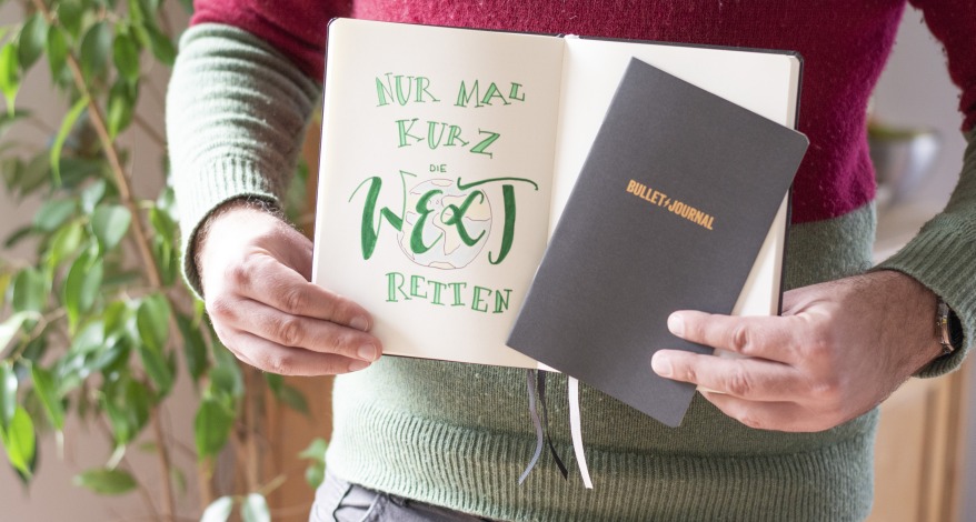 Mann hält Notizbuch mit Lettering „Nur mal kurz die Welt retten“