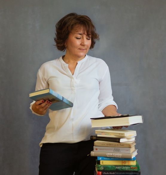 Anja Kuhn steht neben einem Stapel Bücher und hält eins in jeder Hand.