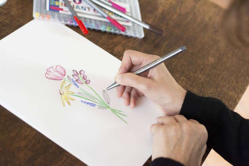 Eine Hand zeichnet auf einem Blatt Papier bei einem gemalten Blumenstrauß mit einem Fineliner von Staedtler Details ein.