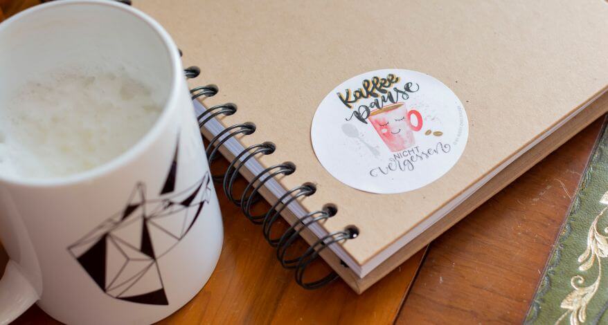 Nahaufnahme von einem Notizbuch, auf dem ein runder Aufkleber von Avery Zweckform klebt. Auf ihm steht in Handlettering: "Kaffeepause nicht vergessen." Daneben steht eine Tasse Milchkaffee.