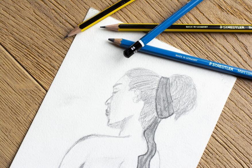 und Die Bleistiftzeichnung einer Frau liegt auf einem Schreibtisch. Um die Zeichnung herum liegen die Bleistifte Noris und Lumograph der Marke Staedtler.
