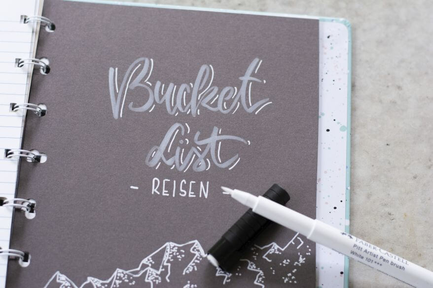 Ein Notizbuch von Filofax liegt aufgeklappt auf einem Schreibtisch, zu sehen ist ein selbstgebastelter Divider, auf dem "Bucket List - Reisen" im Handletteringstil geschrieben steht.
