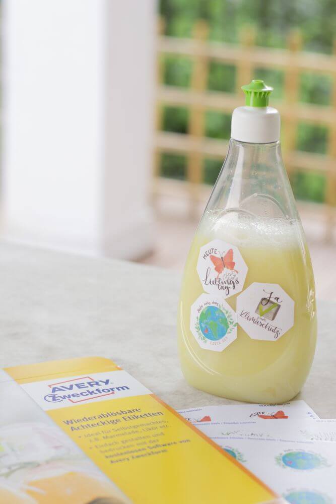 Eine Flasche mit DIY-Spülmittel steht auf einem Tisch. Sie ist beklebt mit achteckigen Aufklebern von Avery Zweckform, der Etikettenbogen liegt daneben.