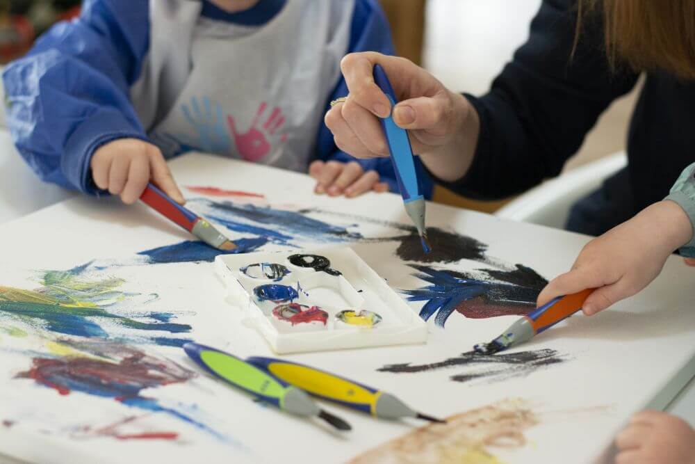 Kinderhände malen mit Pinsel und Goauchefarben aus der Kreativfabrik von Pelikan auf eine Leinwand, ein Erwachsener gibt Hilfestellung.