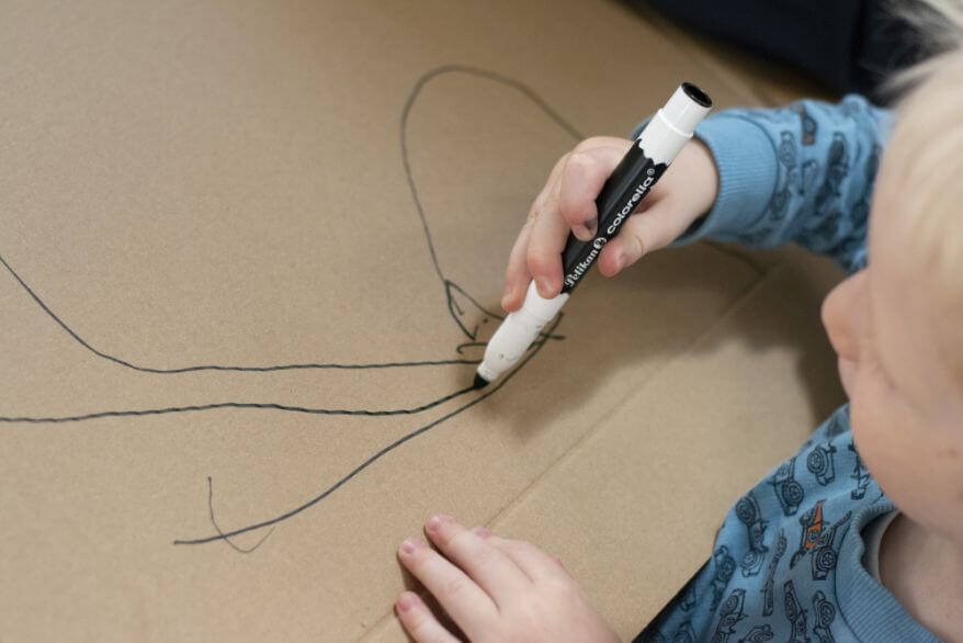 Ein Kind malt mit einem schwarzen Filzstift aus der Kreativfabrik von Pelikan auf ein Stück Pappe.
