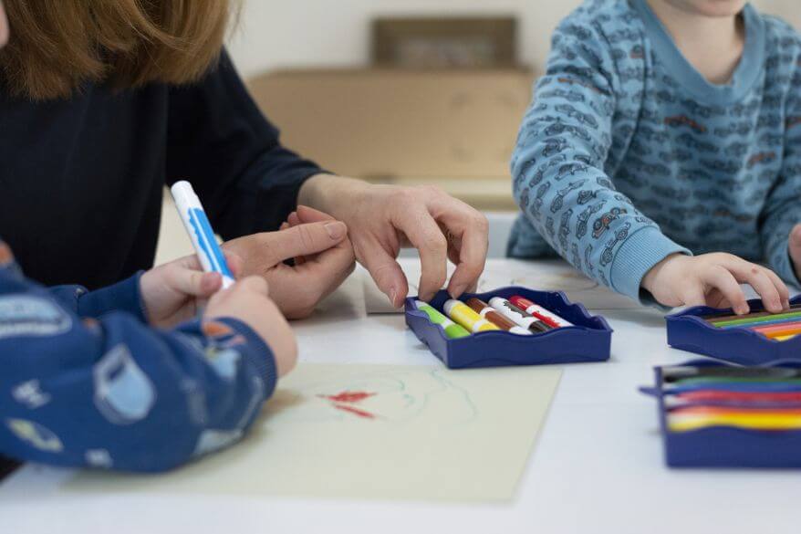 Auf einem Tisch stehen verschiedene Stifte aus der Kreativfabrik von Pelikan, Kinderhände greifen danach.
