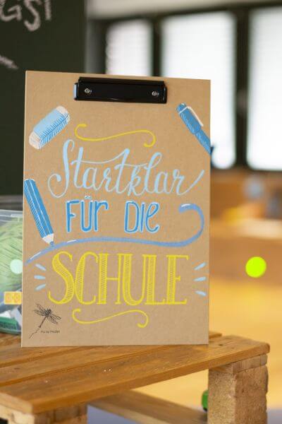 Auf einem Holztisch steht ein Klemmbrett. Dieses wurde mit den Worten "Startklar für die Schule" belettert. Darauf sind außerdem ein Bleistift, ein Füller und ein Radiergummi gemalt. Im Hintergrund ist unscharf eine Tafel zu sehen.