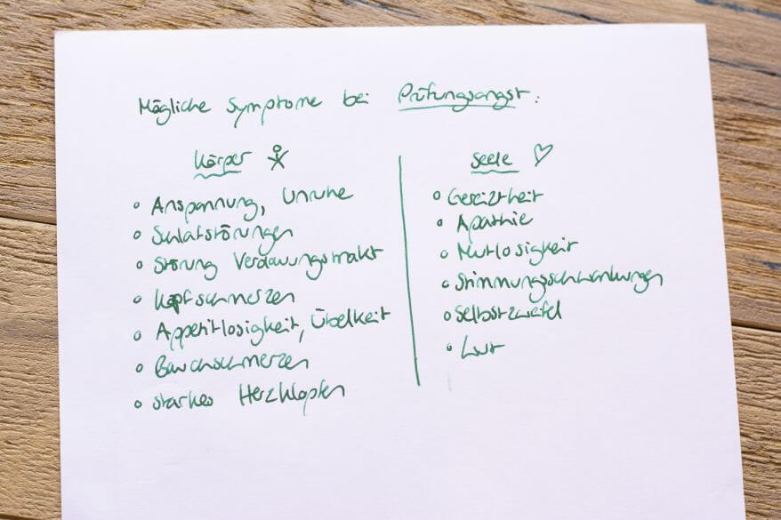 Ein Blatt Papier liegt auf einem Holztisch. Darauf sind mit grüner Tinte mögliche Symptome bei Prüfungsangst aufgelistet. Diese sind auf physische und psychische Symptome aufgeteilt. 