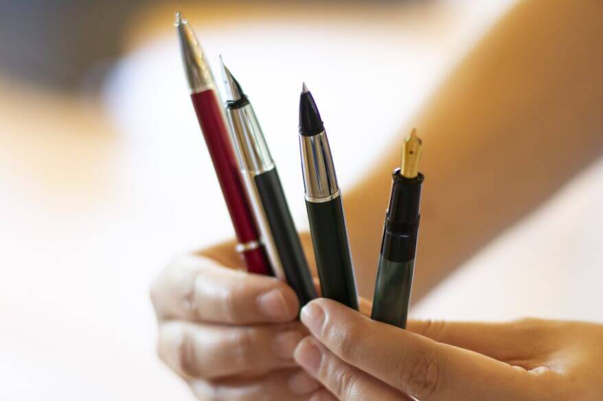 Nahaufnahme verschiedener Schreibgeräte (Füllhalter, Kugelschreiber und Tintenroller) von Pelikan, die aufgefächert von zwei Händen gehalten werden.