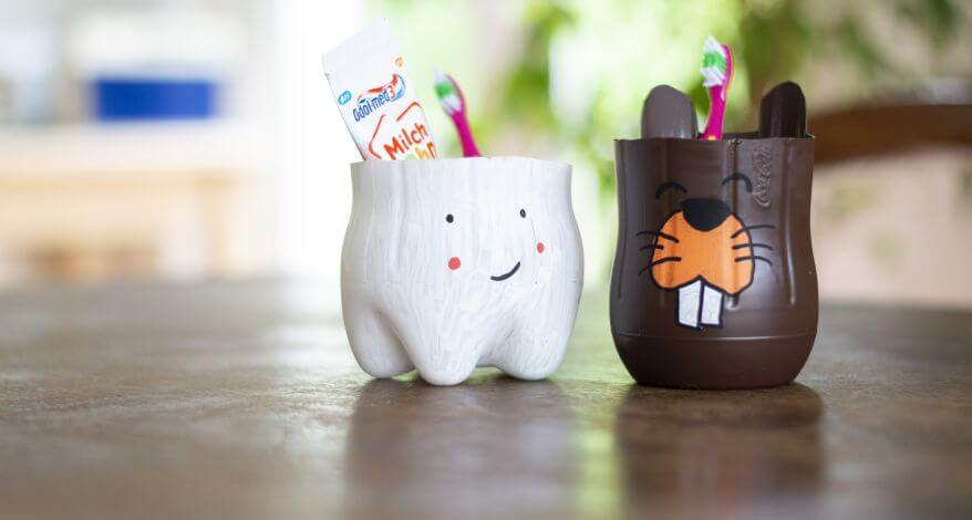 Zwei Zahnputzbecher aus PET-Flaschen im Zahn- & Biebermotiv. In den Bechern stehen Zahnbürsten und Zahnbasta.