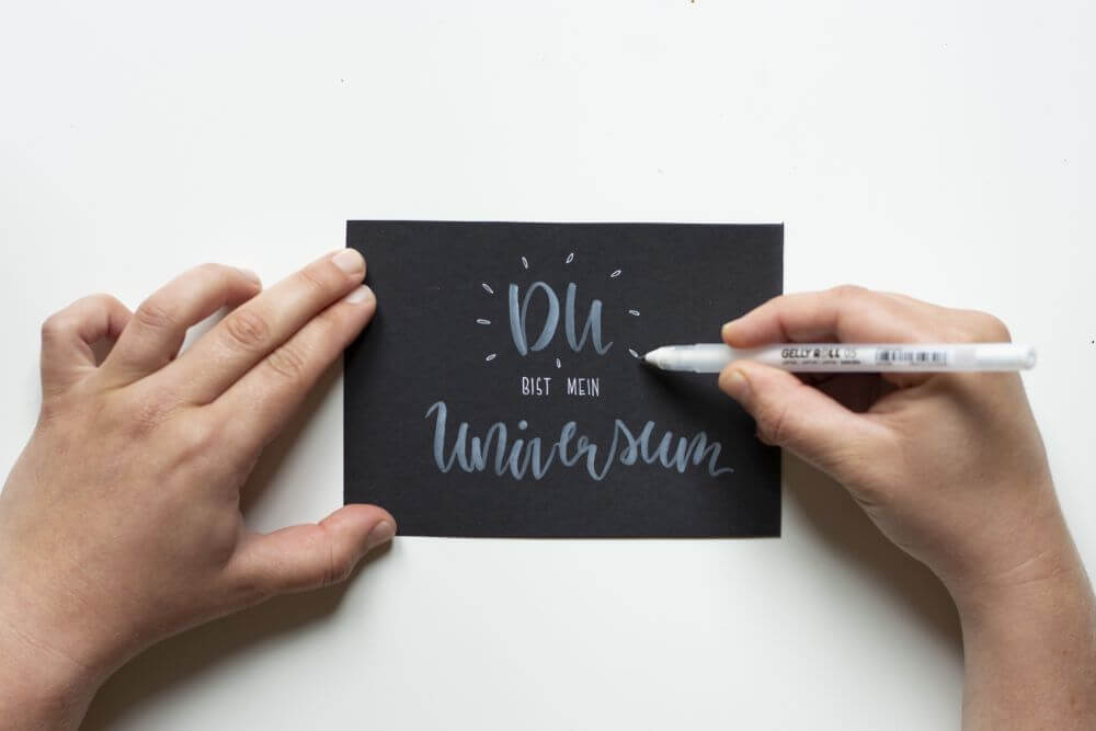 Anleitung Blending mit Gelstiften (Gelly Rolls von Sakura), Schritt 1: Mit weißem Brushpen und weißen Gelroller den Schriftzug "Du bist mein Universum" auf eine schwarze Karte schreiben.