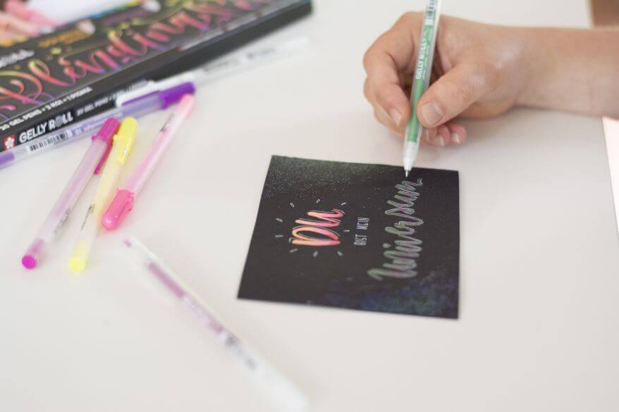 Eine Hand schreibt im Handletteringstil "Du bist mein Universum" auf eine schwarze Karte. Sie verwendet dafür die Gelstifte "Gelly Rolls" von Sakura.