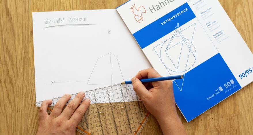 Blick von oben auf einen Schreibtisch, wo zwei Hände eine perspektivische Zeichnung mittels der 3-Punkt-Perspektive auf einem Entwurfsblock von Hahnemühle entwerfen.