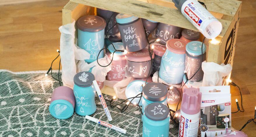 Darstellung eines Upcycling-Adventskalenders aus leeren Marmeladen- und Einmachgläsern: Die Gläser in rosa, grau und türkis wurden in eine Holzkiste gestellt und mit einer Lichterkette dekoriert.