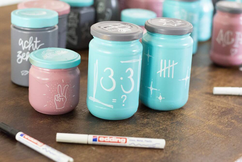 Drei Gläser aus einem Upcycling-Adventskalender aus leeren Marmeladen- und Einmachgläsern stehen auf einem Tisch, davor liegen zwei weiße Glanzlackmarker von Edding.