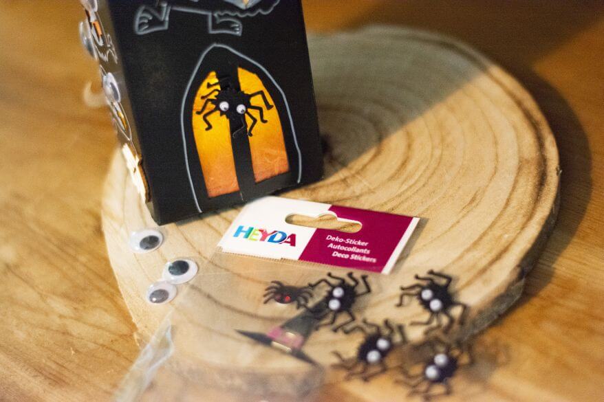 Selbstgemachte Halloweendeko steht in Form eines DIY-Gruselhauses aus einer leeren Milchpackung auf einer Holzscheibe. Darunter liegen Dekosticker in Spinnenform und Wackelaugen von Heyda.