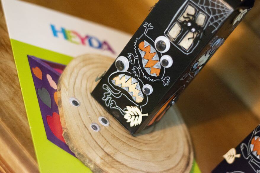 Ein selbstgebasteltes DIY-Gruselhaus zu Halloween aus einer leeren Milchpackung steht auf einer Baumscheibe, darauf liegen Wackelaugen von Heyda, im Hintergrund liegt eine Mappe mit Transparentpapier von Heyda.