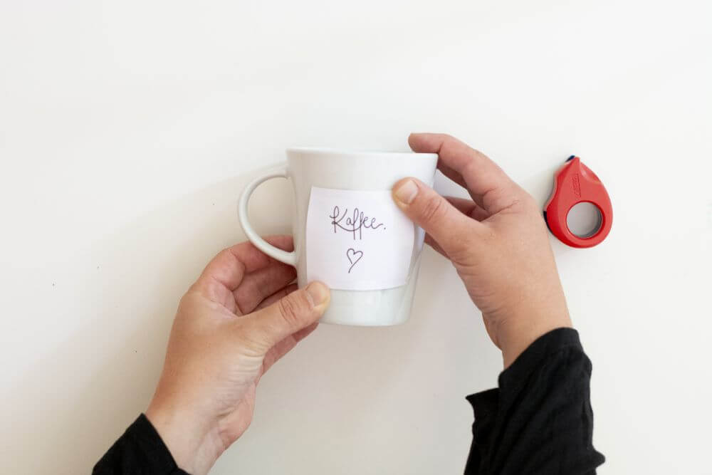Zwei Hände fixieren einen Zettel mit Klebeband auf einer weißen Tasse. Im Hintergrund ist ein Tesafilmabroller in rot zu sehen.
