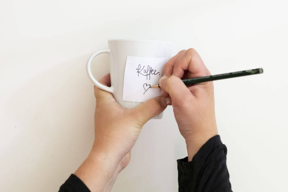 Zwei Hände halten eine weiße Tasse, auf die ein Zettel mit einem Lettering geklebt wurde. Das Lettering und die Zeichnung darunter wird gerade mit einem Bleistift nachgezeichnet. 