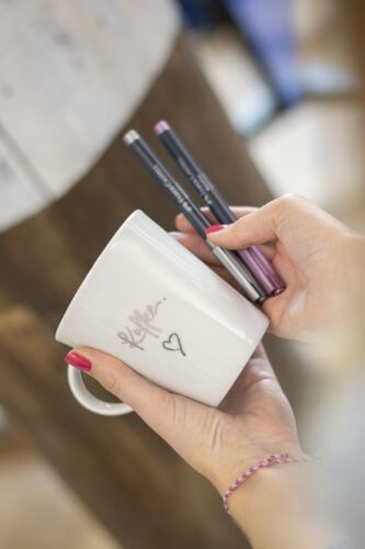 Ein Hand hällt eine beletterte Kaffeetasse, in der anderen Hand liegen Marker von Faber-Castell.