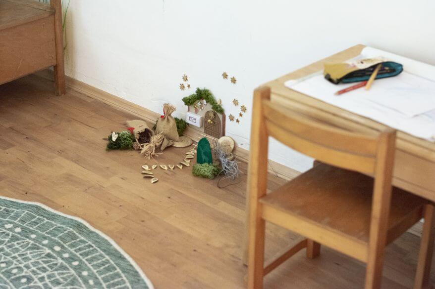 Blick in ein Kinderzimmer: Man sieht einen Maltisch und Kinderstuhl, daneben sieht man sich eine selbstgebastelte Wichteltür an der Wand und auf dem Boden.