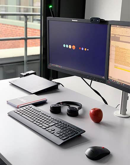 Man sieht einen Arbeitsplatz mit zwei Bildschirmen und einer Webcam. Davor liegen eine Tastatur und eine Maus sowie ein Notibuch, ein Headset sowie ein Apfel.