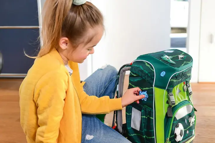 Ein Mädchen in gelber Strickjacke sitzt auf dem Boden vor ihrem grünen Step-by-Step Schulranzen und befestigt mit der linken Hand einen blauen Sticker auf der Seite des Ranzens.