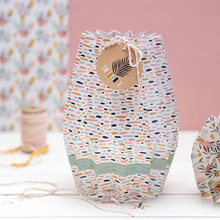 Eine Origami- oder Plissee Vase aus buntem Designpapier der Marke Artoz steht vor einem rosafarbenen Hintergrund. An der Pappvase ist ein bestempeltes Etikett aus Kraftpapier befestigt.