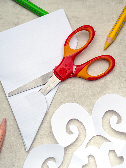 Auf einem grauen Untergrund liegt eine Schulschere der Marke Faber-Castell. Sie schneidet gerade eine Form aus weißem Papier aus. Drumherum sind lose Jumbo Grip Buntstifte verteilt. Foto: Johanna Rundel