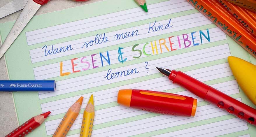 In einem Grundschulheft steht geschrieben "Wann sollte mein Kinde Lesen und Schreiben lernen?". Drumherum liegen Buntstifte der Marke Faber-Castell und ein Schulfüller. Foto: Johanna Rundel