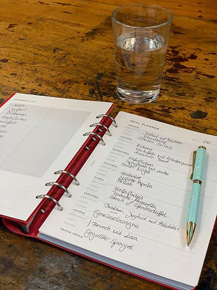 In einem aufgeklappten Notizbuch der Marke Filofax sieht man einen ausgefüllten Mealplanner. Darauf liegt ein geöffneter Kugelschreiber. Am oberen Bildrand erkennt man ein gefülltes Glas mit Wasser. 
