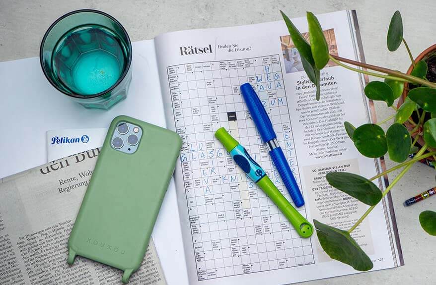 Auf einem grauen Untergrund liegt ein aufgeschlagenes Kreuzworträtsel das zum Teil ausgefüllt ist. Auf dem Rätsel liegen zwei Stifte: Ein Combino Schreiblernfüller und ein griffix Schreiblern-Tintenroller von Pelikan. Auf der linken Seite des aufgeschlagenen Heftes liegen eine Zeitung und ein Smartphone. Außerdem sind ein grünes Wasserglas und eine Zimmerpflanze im Bild zu sehen. Foto: Johanna Rundel 