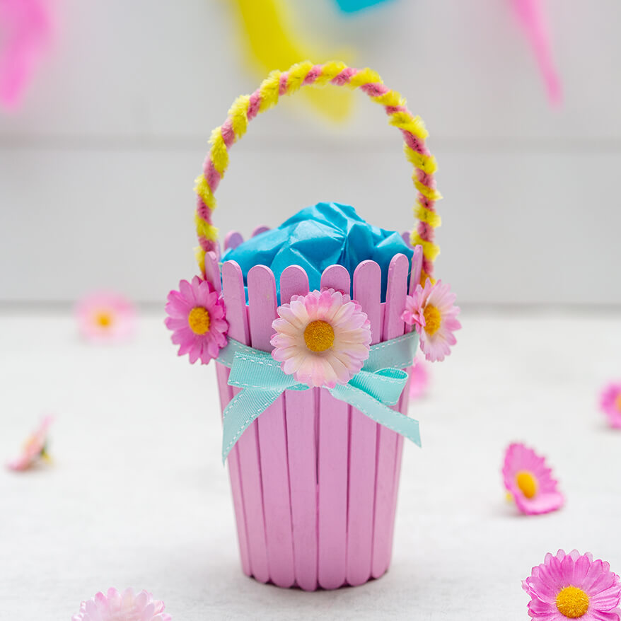 Ein selbstgebasteltes Osterkörbchen, aus einem Pappbecher und Bastelhölzern steht vor einem weißen Hintergrund. Drumherum liegen künstliche Streublumen von Knorr-Prandell. Fotos: Johanna Rundel