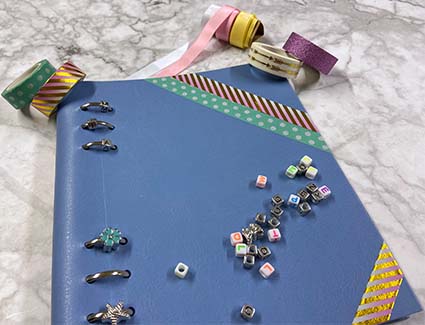Ein Filofax Notizbuch wurde mit Hilfe des Creativekit personalisiert. Um das Buch liegen zahlreiche Washitapes, Charms und farbige Bänder.