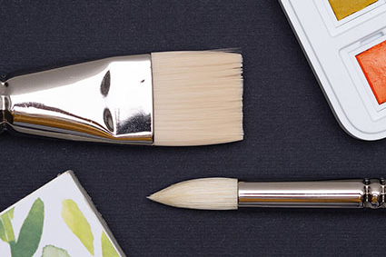 Auf einem schwarzen Untergrund liegen ein Rund- und ein Flachpinsel. Man sieht nur die Spitzen. Rechts oben ist ein Aquarellfarbkasten von Royal Talens zu sehen. In der linken unteren Ecke sieht man einen Aquarellblock von van Gogh der "Frau Hölle-Serie". Foto: Johanna Rundel