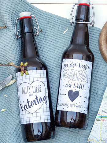 Auf einem blauen Tuch liegen mittig zwei Bierflaschen mit Bügeln. Auf den Flaschen kleben selbstgestaltete Etiketten von Avery Zweckform mit Glückwünschen zum Vatertag in schwarz und weiß. Links neben den Flaschen sind ein paar Zweigen mit frischen, grünen Knospen und ein angespitzter Stock mit einem Taschenmesser zu sehen. Rechts liegen eine aufgefaltete Wanderkarte und ein typischer Wanderstock. Foto: Johanna Rundel