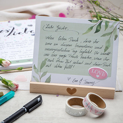 Auf einem hellen Untergrund steht ein Holzaufsteller von Goldbuch mit einem mittig eingebrannten Herz. Darin steckt eine zarte Aquarellkarte auf der die Anleitung für das Hochzeitsgästebuch per Hand geschrieben ist. Im Hintergrund sieht man das aufgeschlagene Hochzeitsgästebuch der Marke Goldbuch. Drumherum liegen verschiedene Stifte, Washi Tapes und Blumen verstreut. Foto: Johanna Rundel