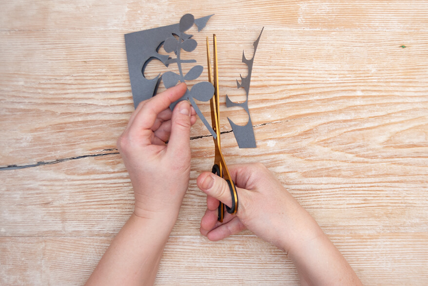 Auf einer hellen Holzoberfläche schneidet jemand mit einer goldenen Schere einen Eukalyptuszweig aus einem dunkelgrauen Blatt Fotokarton von Artoz. Foto: Johanna Rundel