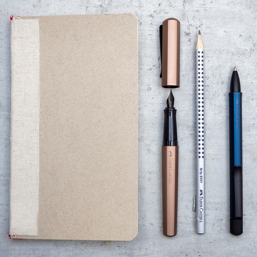 Auf einem Untergrund in Betonoptik liegt links ein beiges Notizbuch. Daneben liegen ein kupferfarbener Hexo Füllfederhalter, ein grauer Grip Bleistift und ein dunkelblauer Ballpoint-Pen der Marke Faber-Castell. Foto: Johanna Rundel
