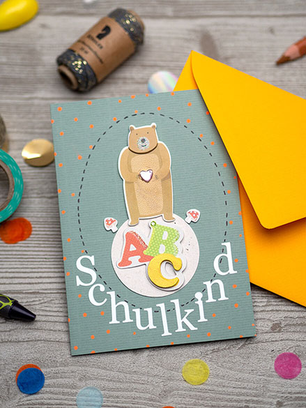 Auf einem hellgrauen Holzuntergrund liegt eine wasserblaue Grußkarte auf die ein brauner Bär, die Buchstaben ABC und das Wort Schulkind mit Aufklebern der Firma Rössler geklebt ist. Drumherum liegen buntes Konfetti, eine Garnspule, Washitape und verschiedene Stifte verteilt.